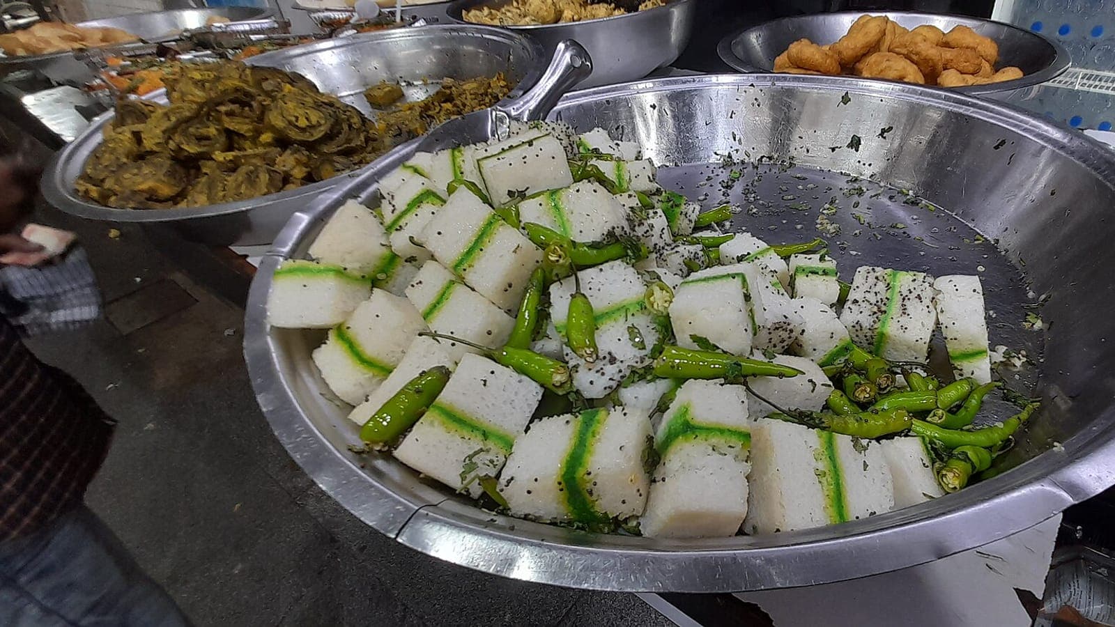 Trays of yellow dhokla and fried snacks laid out in steel dishes at a food counter